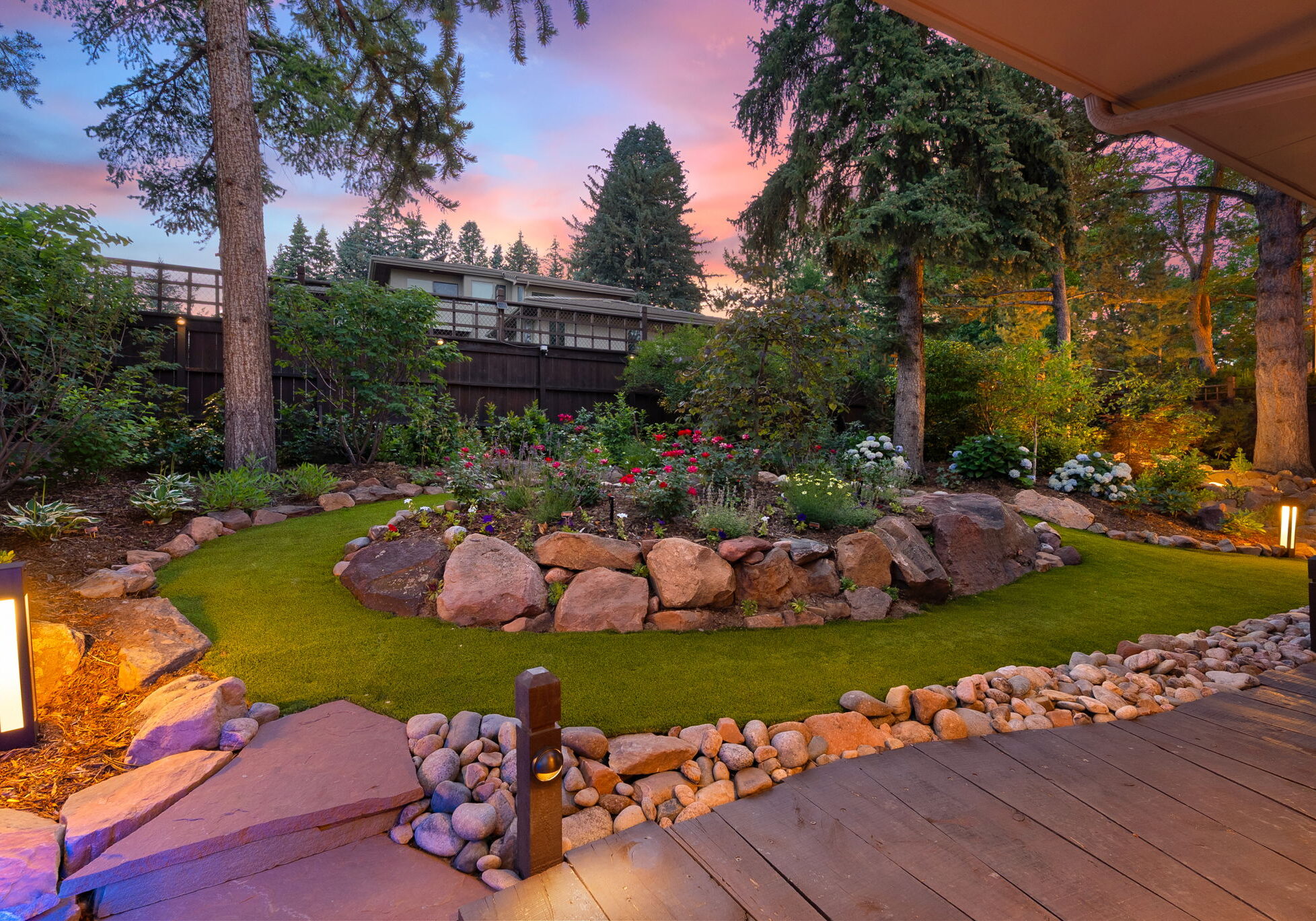 Manicured lawn island with blooming rose and perennial garden beds, boulder border, and glowing bollard lights at twilight – Boulder County landscape maintenance by Nature's Way Landscapes
